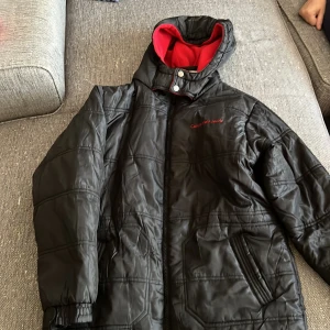 Svart jacka från Calvin Klein Jeans - Säljer en svart Calvin Klein Jeans jacka i storlek L. Den har en röd insida och en huva för extra värme. Jackan är perfekt för kyligare dagar och har en dragkedja framtill. Den är i bra skick och redo för nya äventyr!