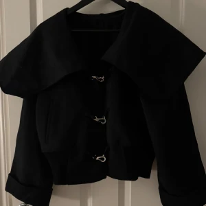 Svart kappa med spännen - Säljer en stilren svart kappa med spännen framtill. Den har en stor krage och ribbad nederkant för en snygg passform. Storlek S