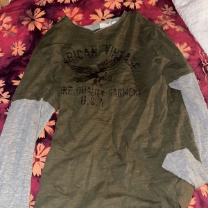 Grön långärmad tröja från Kappahl - Säljer en cool grön tröja från Kappahl med tryck av en örn och texten 'American Vintage'. Tröjan har långa grå ärmar som ger en lager-på-lager-look. Perfekt för höst och vår! 🦅