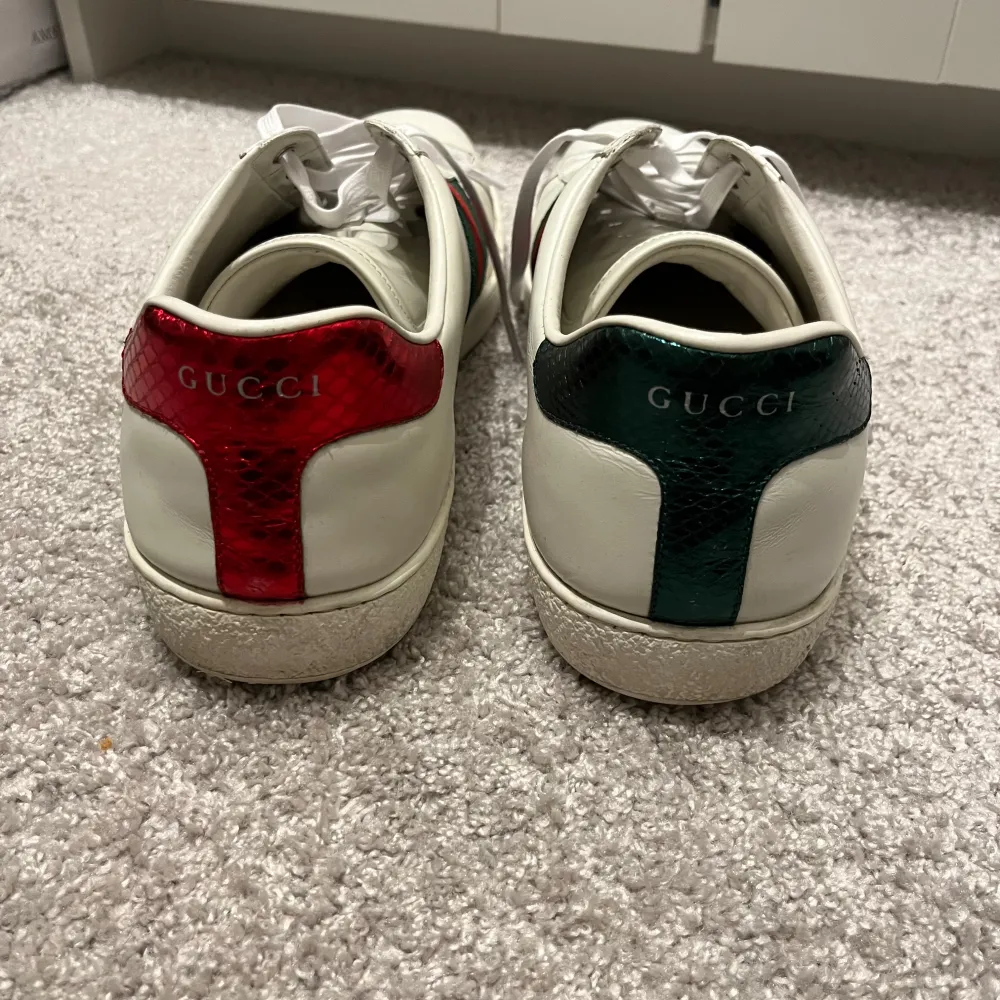 Säljer ett par vita Gucci Ace sneakers med klassiska gröna och röda ränder samt ett broderat emblem på sidan. Skorna är i bra skick med helt nya skosnören och har lite slitage på innersulan. Perfekta för både vardag och fest!. Skor.