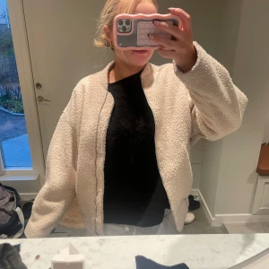 Beige teddyjacka - Mysig beige teddyjacka i oversized stil. Perfekt för höst och vinter med sin fluffiga och varma design. Jackan har långa ärmar och en dragkedja framtill. Den är både stilren och bekväm, idealisk för kalla dagar.