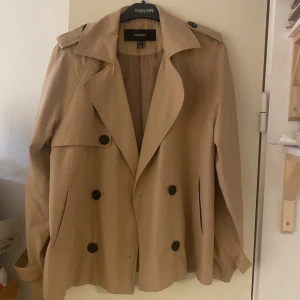 Beige trenchcoat från Vero Moda - Säljer en snygg beige trenchcoat från veromoda i mycket bra skick. Jackan har en klassisk design med dubbelknäppning och stora knappar. Perfekt för höst och vår, med långa ärmar och stiliga detaljer vid axlarna. Passar både till vardags och lite finare tillfällen. Strl M