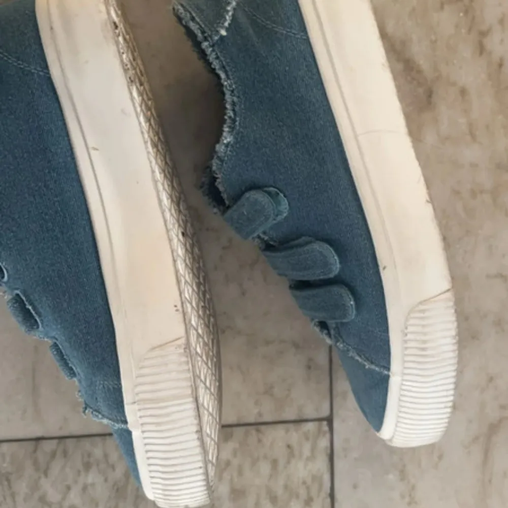 Säljer ett par coola blå sneakers från EVEN&ODD i jeansmaterial. De har tre praktiska kardborreband och en vit sula. Perfekta för en avslappnad stil och passar till både vår och sommar. Skorna har en fransig kant som ger en extra touch av stil.. Skor.