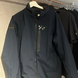 Mörkblå jacka från Helly Hansen - Säljer en mörkblå jacka från Helly Hansen i mycket bra skick. Jackan har en dragkedja framtill och är perfekt för höst och vår. Den är tillverkad i syntetmaterial och har en normal passform. Perfekt för dig som vill ha en stilren och funktionell jacka!