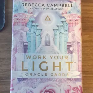 Säljer en vacker kortlek 'Work Your Light Oracle Cards' av Rebecca Campbell med konst av Danielle Noel. Förpackningen har ett drömskt och färgglatt omslag med rosa och blå nyanser samt mystiska symboler och blommor. Perfekt för den som är intresserad av andlighet och personlig utveckling.