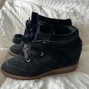 Svarta Isabel Marant - Säljer ett par snygga svarta sneakers från Isabel Marant i storlek 39. De är gjorda i mocka och har en cool kilklack för extra höjd. Skorna har snörning framtill och en bekväm gummisula. Perfekta för både vardag och fest!