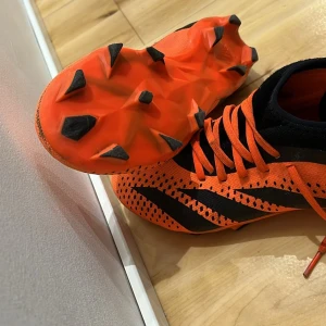 Orange och svarta fotbollsskor - Säljer ett par grymma fotbollsskor i orange och svart. De har en snygg design utan snörning och är perfekta för spel på gräs. Skorna är i bra skick och redo för nästa match! Jag har tagit bort skosnören!