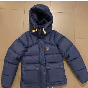 Blå dunjacka från Fjällräven - Säljer en snygg blå dunjacka från Fjällräven. Den har en mysig huva och gula detaljer vid snörningen. Perfekt för kalla vinterdagar! Jackan har knappar och dragkedja framtill för extra skydd mot kylan. Passar både till vardags och utomhusaktiviteter. Den har en liten defekt vid armen men annars helt som ny därför jag säljer för billigare pris