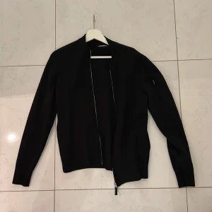 Svart jacka från J.Lindeberg - Säljer en stilren svart kofta från J.Lindeberg i storlek S. Jackan har en dragkedja framtill och är långärmad. Perfekt för höst och vår, och passar både till vardags och fest. Den är i bra skick och redo för en ny ägare, den är endast lite nopprig men går att få bort 