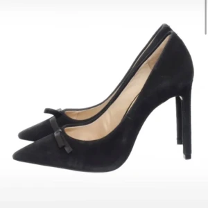 Svarta pumps med rosett - Snygga svarta pumps i mocka med spetsig tå och elegant rosett på framsidan. Perfekta för festliga tillfällen eller en utekväll. De har en hög klack som ger en stilren look. Skorna är i mycket bra skick och redo att användas för att lyfta din outfit!