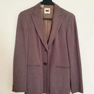 Brun kavaj från DKNY - Snygg brun kavaj från DKNY i petite-storlek. Perfekt för både kontoret och mer formella tillfällen. Kavajen har en klassisk design med två knappar framtill och fickor. Materialet känns lyxigt och ger en stilren look. Passar perfekt till höst- och vårgarderoben.