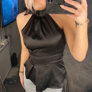Svart fest topp - Snygg svart topp i satin med en elegant glans. Toppen har en åtsittande passform i midjan och en lätt utsvängd nederdel. Perfekt för en utekväll eller fest! 🖤