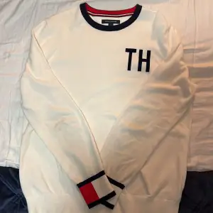 Säljer en snygg vit tröja från Tommy Hilfiger med 'TH' broderat på bröstet. Den har långa ärmar och ribbade muddar med röd och marinblå detalj. Perfekt för en stilren och casual look. Passar både till vardags och fest! Denna tröja kommer i storlek M! Den är i ett mycket bra skick 10/10!