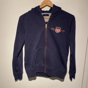 Mörkblå hoodie från GANT - Säljer en snygg mörkblå hoodie från GANT i storlek 146/152 cm, perfekt för 11-12 åringar. Den har en dragkedja framtill och ett coolt GANT-emblem på bröstet. Tröjan är i bra skick och passar perfekt för höst och vår. Skön och stilren design som funkar till det mesta!