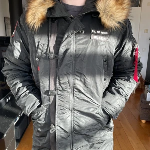 Svart alpha industries vinterjacka - Svart fodrad vinterjacka från alpha industries med fejkpälskrage. Röda och vita detaljer. Fickor på framsida och insida, två på magen, två på bröstet och en på insidan. Dragkedje och knapp förslutning. Går strax under rumpan (jag är 188cm). Storlek L
