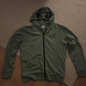 Grön hoodie från C.P. Company - Säljer en grön hoodie från C.P. Company i storlek 14 år. Den har en unik design med inbyggda glasögon i huvan och dragkedja framtill. Perfekt för höst och vår, och ger en cool look till vardagsoutfiten. Använd ett fåtal gånger, så den är i mycket bra skick. Passar perfekt för dig som gillar streetwear!