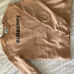 Beige tröja från Burberry - Säljer en snygg beige tröja från Burberry i storlek S. Den har en klassisk design med Burberry-loggan tryckt i svart på framsidan. Tröjan är i bomullsmaterial och har ribbade muddar vid ärmslut och nederkant. Perfekt för en stilren och avslappnad look!