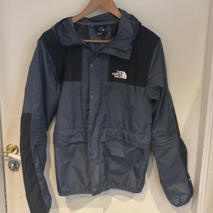 Grå och svart jacka från The North Face - Säljer en grå och svart jacka från The North Face i mycket bra skick. Jackan har en huva och långa ärmar, perfekt för höst och vår. Den är tillverkad i ett slitstarkt syntetmaterial och har en dragkedja framtill. Passar bra för utomhusaktiviteter och vardagsbruk.