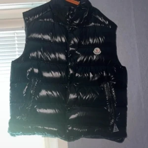 Moncler väst - Säljer en snygg svart dunväst från Moncler. Västen har en glansig yta och är försedd med Monclers ikoniska logotyp på bröstet. Perfekt för kyligare dagar när du vill hålla stilen. Västen har praktiska fickor med dragkedja. Säljer den pga liten storlek!! Kan gå ner i pris för den:) bild kvaliten är skit som ni kan se!