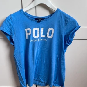 Blå t-shirt från Ralph Lauren - Säljer en blå t-shirt från Ralph Lauren med vit text 'POLO' på framsidan. T-shirten har en rund hals och korta ärmar. Perfekt för en avslappnad stil. Passar bra till jeans eller shorts.