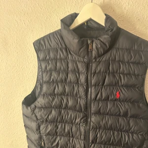 Mörkblå dunväst från Ralph Lauren - Snygg mörkblå dunväst från Ralph Lauren med broderad logga i rött på bröstet. Perfekt för kyligare dagar men passar också til våren. Använd fåtal gånger