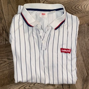 Vit randig pikétröja från Levi's - Säljer en snygg vit pikétröja från Levi's med blå ränder och röd detalj vid kragen. Tröjan har en klassisk pikékrage med knappar och Levi's-logga på bröstet. Perfekt för en stilren look!