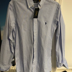 Blå och vit randig Ralph Lauren skjorta - Säljer en helt ny oanvänd blå och vit randig skjorta från Ralph Lauren. Skjortan har långa ärmar och knappar framtill. Den har den ikoniska loggan i blått broderad på bröstet. Storlek XS. Ny pris: 1800! Mitt Pris: 1200! Perfekt för våren och sommaren. 