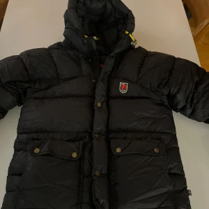 Svart dunjacka från Fjällräven - Jättefin fjällräven jacka. Tyvärr upptäckte jag att jackan fått ett hål på baksidan av armen nu när jag ska sälja. Hålet var som man ser på bilden 2/ 2,5cm. Jackan är i väldigt bra skick i storlek xxxs som motsvarar xs/s. Som också finns med på bild är det en liten sminkfläck på insidan av luvan, inget som syns när jackan är på eller om man inte kollar extra noggrant. Skriv för ännu fler bilder!☺️