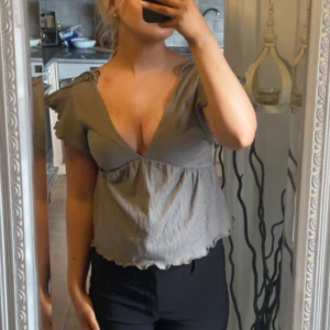 Grå/brun med volangdetaljer - Säljer en söt grå/brun blus med volangdetaljer och djup v-ringning. Blusen har korta ärmar och en luftig passform, perfekt för varmare dagar. Och öppen med knyte i ryggen ( bilden jag tog är snörena uppdragna❤️