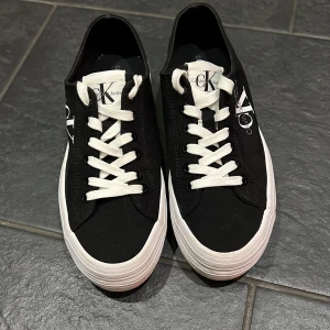 Svarta sneakers från Calvin Klein - Snygga svarta sneakers från Calvin Klein med vit sula och vita snören. Skorna har en klassisk design med märkets logotyp tryckt på sidan. Perfekta för en stilren look. Nästan helt oanvända och i väldigt bra skick