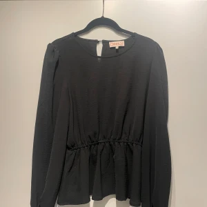 Svart blus från ONLY - Snygg svart blus från ONLY med långärmad design och en lätt rynkad midja för en smickrande passform. Perfekt för både vardag och speciella tillfällen. Blusen har en enkel knäppning i nacken.