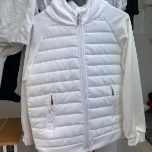 Vit quiltad jacka med huva, cardigan - Säljer en stilren vit quiltad jacka/cardigan med dragkedja och huva. Jackan har långa ärmar och två praktiska fickor med dragkedjor framtill. Perfekt för kyligare dagar. Aldrig använd. 10/10 storlek M. BVSA.
