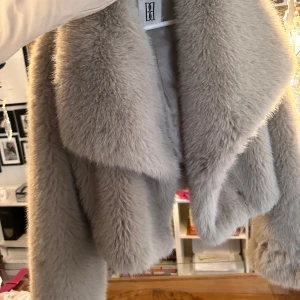 Grå pälsjacka cropped med pälsmössa - Snygg och fluffig cropped grå pälsjacka med mössa till som ger en lyxig känsla. Perfekt för att hålla värmen under kalla dagar. Pälsen är fuskpäls och har snygga platta dubbel knäoäppta tryckknappar för stängning. 
