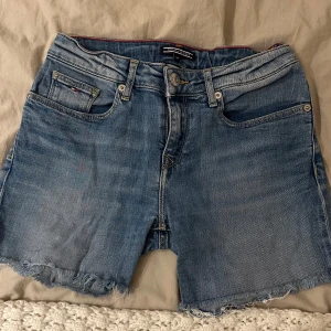 Blå jeansshorts från Tommy Hilfiger - Snygga blå jeansshorts från Tommy Hilfiger med fransiga kanter och klassisk femficksdesign. Perfekta för en avslappnad sommarlook. De har en knapp och dragkedja framtill samt bälteshällor.Storlek 164/s/34