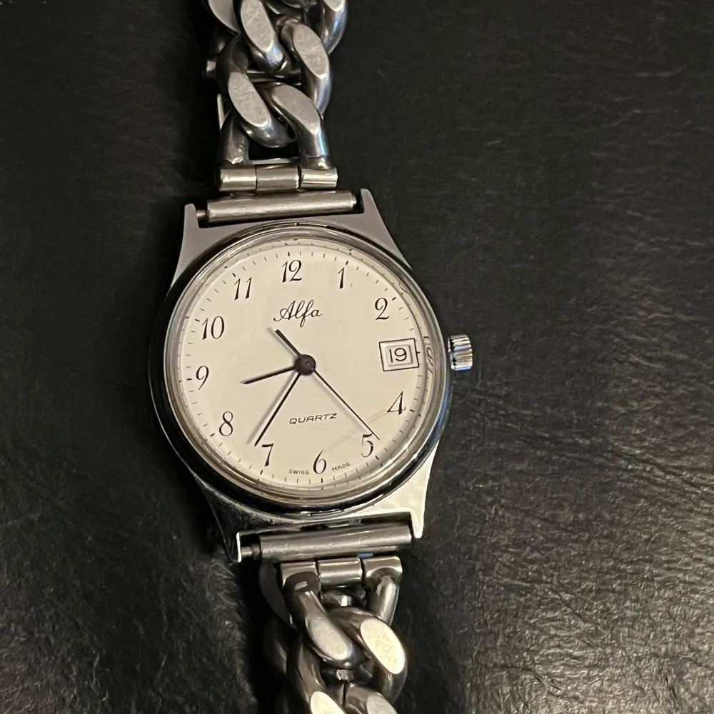 Säljer en stilren silverfärgad klocka från Alfa med vit urtavla och svarta siffror. Klockan har ett datumfönster vid klockan tre och är Swiss Made. Armbandet är i rostfritt stål med en robust länkdesign.. Asusteet.