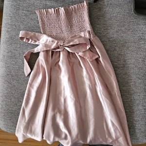 Ljusrosa axelbandslös klänning - Säljer en söt ljusrosa axelbandslös klänning med en stor rosett i midjan. Klänningen har en smockad överdel och ett flödande kjolparti. Perfekt för sommarens festligheter!