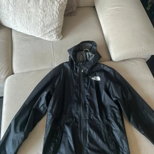 Svart vindjacka från The North Face - Säljer en svart vindjacka från The North Face i storlek L/G för ungdomar. Jackan har en praktisk huva och dragkedja framtill. Perfekt för blåsiga dagar och enkel att packa med sig. 🖤