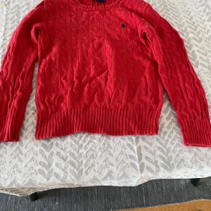 Röd stickad tröja från Ralph Lauren - Säljer en snygg röd stickad tröja från Ralph Lauren med kabelstickat mönster och den klassiska loggan broderad på bröstet. Tröjan har långa ärmar och ribbade muddar vid ärmslut och nederkant. Perfekt för kyligare dagar!