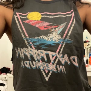 Två ärmlösa toppar med tryck - Säljer två ärmlösa toppar. Den ena är mörkgrå med ett färgglatt tryck av en surfare och texten 'Daytrippin in Bermuda'. Den andra är grå och enkel utan tryck. Perfekta för en avslappnad stil. Kan även köpas styckvis för 40kr/styck