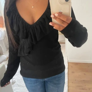 Svart tröja med volangdetalj - Snygg svart tröja med volangdetalj framtill. Tröjan har långa ärmar och en djup v-ringning som ger en elegant touch. Perfekt för att klä upp en vardagsoutfit eller för en kväll ute.