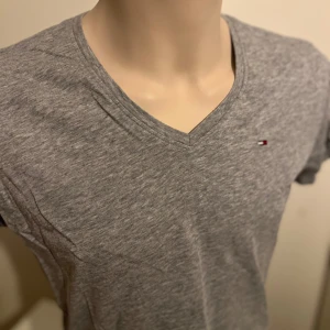 Grå t-shirt från Tommy Jeans - Snygg grå t-shirt från Tommy Jeans med v-ringning och liten logga på bröstet. Perfekt för en avslappnad stil. Tillverkad i mjuk bomull för extra komfort.