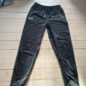 Svarta träningsbyxor från Under Armour - Säljer ett par svarta träningsbyxor från Under Armour. Byxorna har en loose passform med snörning i midjan och dragkedjor vid fickorna. Perfekta för träning eller en avslappnad dag.
