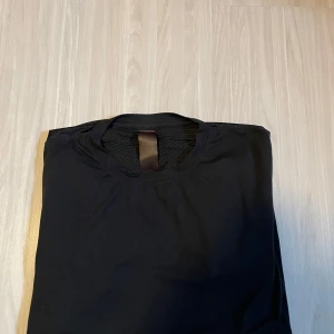 Svart tränings T-shirt från NewBody - Säljer en stilren svart t-shirt med rund hals. Perfekt för en enkel och klassisk look. T-shirten är kortärmad och tillverkad i ett mjukt material.