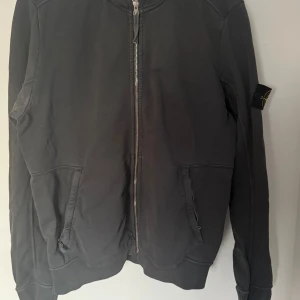 Grå zip-hoddie från Stone Island - Säljer en svart huvtröja från Stone Island med dragkedja framtill. Tröjan har långa ärmar och en klassisk huva. Perfekt för en stilren och avslappnad look.