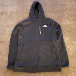 Grå hoodie från The North Face 13-14 - Säljer en svart hoodie från The North Face med dragkedja och huva. Jackan har en bröstficka med dragkedja och två sidofickor. Perfekt för kyliga dagar och har en stilren design med logotyp på bröstet och ryggen. Storleken är cirka 164.