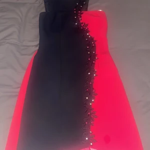 Svart och röd klänning från Erkul - Snygg svart och röd klänning från Erkul med dekorativa blommor och pärlor längs kanten. Klänningen har en dragkedja bak och smockdetaljer vid bysten för en bekväm passform. Perfekt för speciella tillfällen!