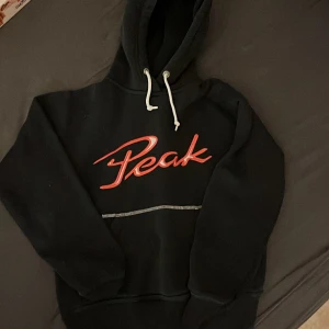 Svart hoodie från Peak - Säljer en svart hoodie från Peak med röd logga på framsidan och siffran 05 på baksidan. Den har en klassisk design med huva och dragsnören. Perfekt för en avslappnad stil. I storleken M men skulle säga att den är xxs-xs
