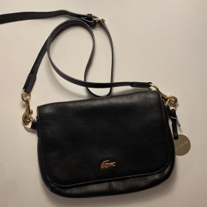 Svart axelväska från Lacoste - Beautiful black crossbody bag with gold details from Lacoste. Looks perfect from outside but a little worn inside, as you can see in the last picture there are a few lipstick and pen stains. Hence some discount can be offered 🖤