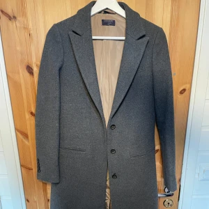 Grå varm kappa från Crocker Stockholm - Snygg grå kappa från Crocker med klassisk design. Den har en stilren look med knappar framtill och långa ärmar. Perfekt för kyligare dagar och ger en elegant touch till din outfit.