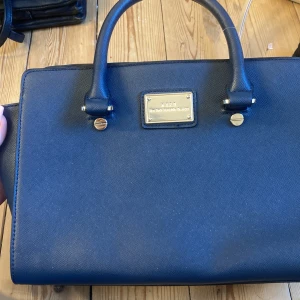 Blå handväska från NYPD - Snygg blå handväska från NYPD med stilren design och metallplatta framtill. Väskan har ett ljust, mönstrat innerfoder och praktisk dragkedja för säker förvaring. Perfekt för att bära med sig det viktigaste med stil.